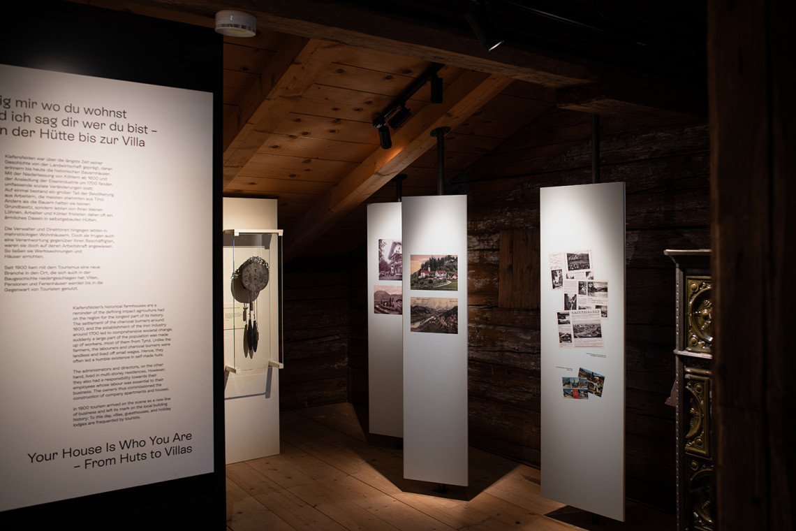 Exhibition view with triangular rotating panels in the ‘Living’ section and two original objects from Kiefersfelden villas: a clock and a fir-green tiled stove.