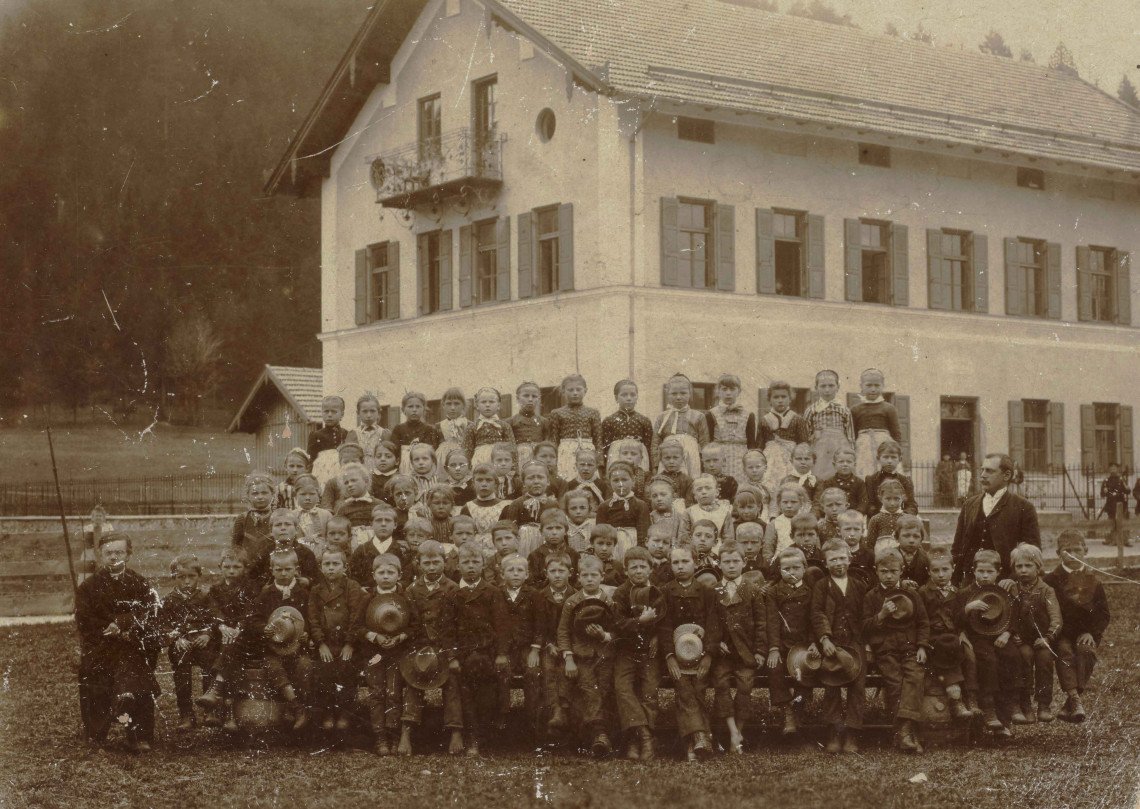 A sepia-coloured group photo showing at least 78 children and two male teachers standing in front of a newly constructed rural, three-storey school building.